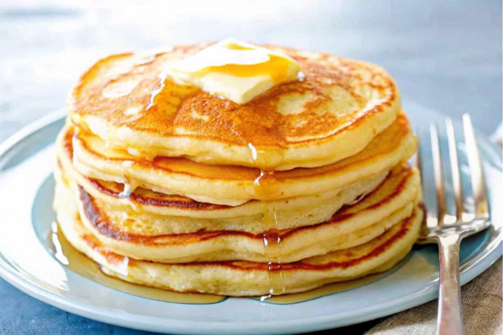 A stack of fluffy, golden Good Old-Fashioned Pancakes topped with fresh berries and maple syrup, served with whipped cream in a cozy breakfast setting.